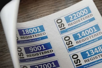 Selos e certificados de registro visíveis em etiquetas com números 9001, 14001, 22000 e 18001, em fundo de papel e textura de madeira, associados à gestão e conformidade industrial. Foto ilustrativa para NR-12: Adequação e Segurança Industrial.