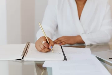 Profissional em ambiente de escritório escreve anotações em um documento aberto com caneta dourada, enquanto organiza tarefas em uma rotina profissional. O papel da organização e planejamento na rotina profissional é evidenciado pelo foco em planejamento e registro.